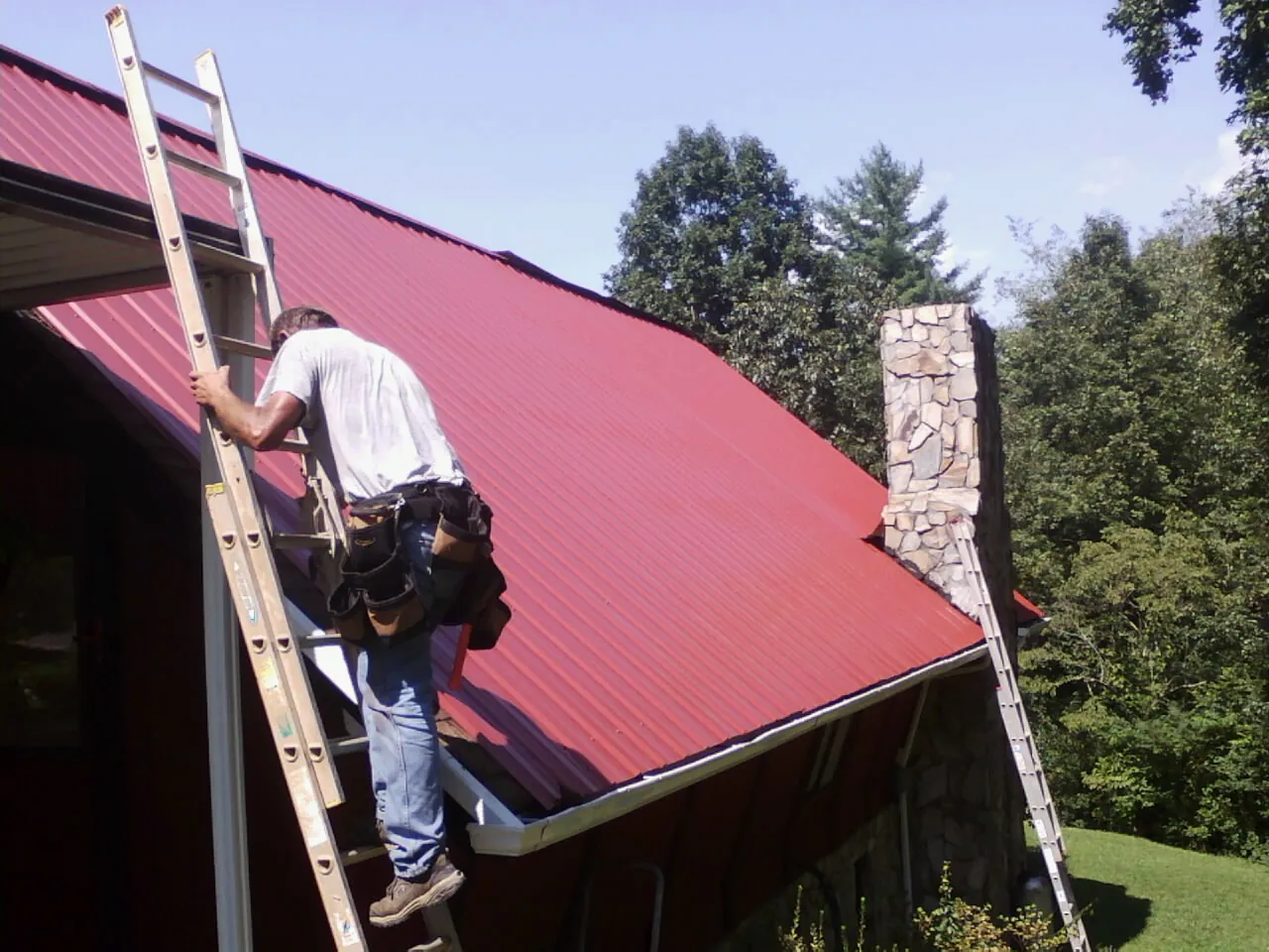 Roofer on a ladder beside a red metal roof and stone chimney — Soffit & Fascia Repair by Roofing Brothers in Pawcatuck
